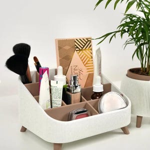Peut inclure: Un organisateur de maquillage blanc et marron avec plusieurs compartiments, contenant divers produits de beauté tels que des pinceaux, des fards à paupières et des produits de soin. L'organisateur a une texture côtelée et repose sur de petits pieds. Une plante en pot est en arrière-plan.