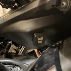 May include: Close-up of a motorcycle's black fairing, featuring a small, rectangular black control module with two silver buttons. The module is mounted on the fairing, with various mechanical components visible in the background.