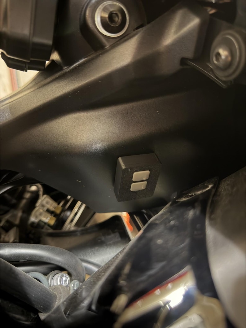 May include: Close-up of a motorcycle's black fairing, featuring a small, rectangular black control module with two silver buttons. The module is mounted on the fairing, with various mechanical components visible in the background.