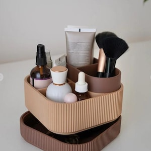 May include: A two-tiered cosmetic organiser in beige and brown. The top tier holds beauty products like bottles, tubes, and brushes. The bottom tier is a tray. The ribbed design adds texture. The organiser is designed to keep beauty essentials tidy and organised.