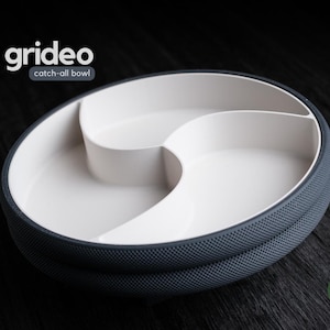 Può includere: Una moderna ciotola multiuso con interno bianco ed esterno strutturato grigio scuro. La ciotola è divisa in tre sezioni. Le parole "grideo" e "catch-all bowl" sono stampate in alto a sinistra.