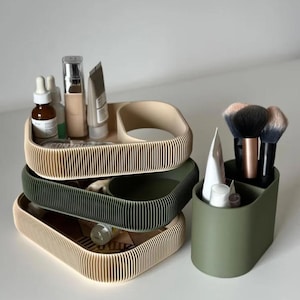 Peut inclure: Trois plateaux cosmétiques côtelés beiges et verts empilés, et un porte-pinceaux vert assorti. Les plateaux contiennent des produits de beauté. Le porte-pinceaux contient des pinceaux de maquillage.