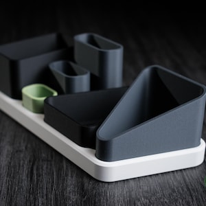 Peut inclure: Un ensemble d'organisateur de bureau moderne dans des tons de gris, noir et vert. L'ensemble comprend divers contenants de formes variées, tels que rectangulaires, triangulaires et ovales, disposés sur une base rectangulaire blanche. Le design est minimaliste et fonctionnel.