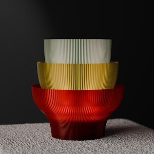May include: Three decorative bowls stacked on top of each other, in red, gold, and pale green. Each bowl has a ribbed texture. The bowls are on a white textured surface against a dark background.