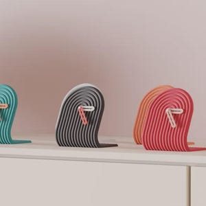 May include: Three decorative bookends in teal, grey, and red-orange, each with a wave design. A white paperclip is attached to each bookend. A stack of books is to the right, all on a white surface against a pink background.