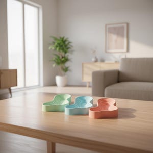 Op de afbeelding: Drie pastelkleurige, golfvormige decoratieve trays staan op een houten salontafel. De trays zijn lichtgroen, lichtblauw en koraal. De trays hebben een gelaagd ontwerp en staan in een moderne woonkamer.