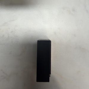 May include: A rectangular black object, possibly a small box or case, is positioned vertically against a light-colored surface. The object appears to be solid and opaque, with a smooth, dark finish. A small, silver-colored detail is visible on the side.