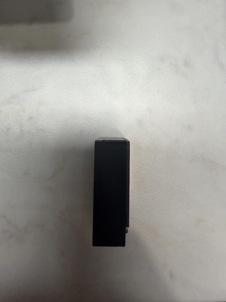 May include: A rectangular black object, possibly a small box or case, is positioned vertically against a light-colored surface. The object appears to be solid and opaque, with a smooth, dark finish. A small, silver-colored detail is visible on the side.