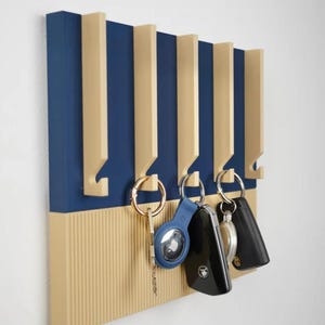 Peut inclure: Un porte-clés mural moderne, bleu marine et beige. Il présente des fentes verticales pour accrocher les clés, avec plusieurs jeux de clés suspendus aux crochets. Le design est minimaliste et contemporain, idéal pour la maison.
