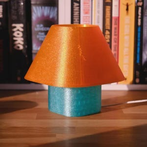 May include: A small decorative lamp with a teal base and an orange lampshade. The lampshade is a trapezoid shape. The lamp is on a wooden surface in front of a bookshelf filled with books.