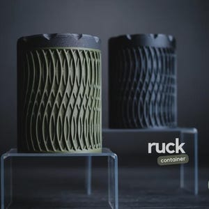 May include: Two cylindrical containers with textured wave patterns. One is olive green, the other black. Both have dark grey lids and sit on clear acrylic stands. The word "ruck" and "container" are visible.