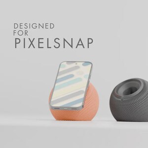 Könnte beinhalten: Ein Smartphone steht in einem orangefarbenen, kugelförmigen Ständer neben einem grauen, kugelförmigen Lautsprecher. Der Bildschirm des Telefons zeigt ein farbenfrohes Muster. Der Text "DESIGNED FOR PIXELSAP" steht oben.