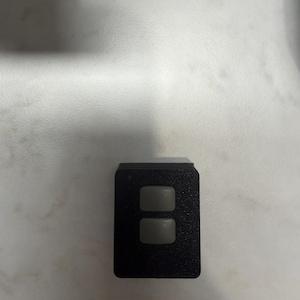 May include: A small, rectangular black device with two gray, square buttons. The device has a textured surface and is likely a remote control or a similar electronic component.