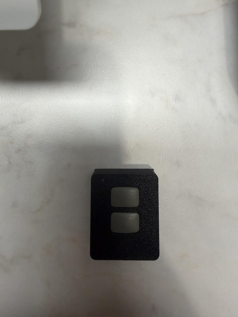 May include: A small, rectangular black device with two gray, square buttons. The device has a textured surface and is likely a remote control or a similar electronic component.