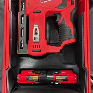 May include: A red Milwaukee cordless nail gun with a black handle and a metal belt clip, stored inside a red hard-shell carrying case with a custom black insert. Two red and black battery packs are positioned in the lower section of the case.