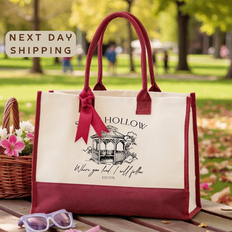A red and white tote bag with a graphic design and text, placed on a wooden surface with a basket of flowers and sunglasses nearby.