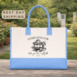 A blue and white tote bag with the text "STARS HULLOW" and "Where you have loved I will follow" printed on it, standing on a wooden surface with a garden in the background.