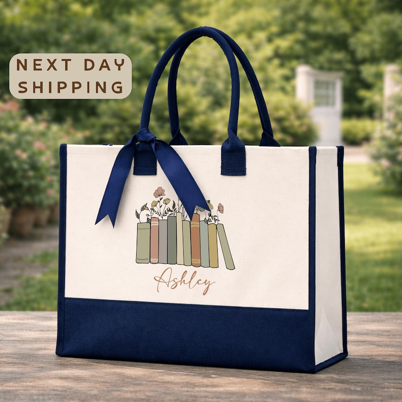 A white tote bag with a blue ribbon and a graphic of a book, placed on a wooden surface with a garden in the background.