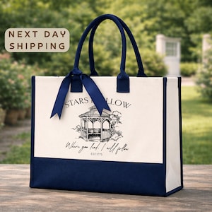 A white tote bag with a blue ribbon and a graphic of a gazebo, placed on a wooden surface against a blurred background of greenery.