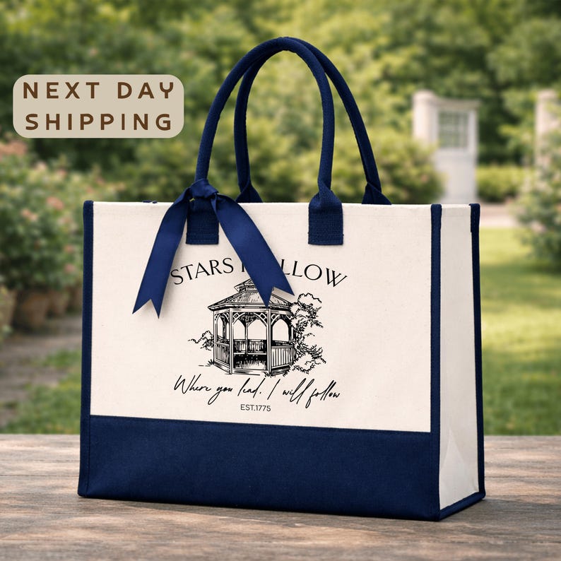 A white tote bag with a blue ribbon and a graphic of a gazebo, placed on a wooden surface against a blurred background of greenery.