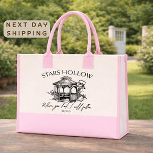 A pink tote bag with a printed design of a gazebo and the text "STARS HOLLOW" printed on it, placed on a wooden surface.