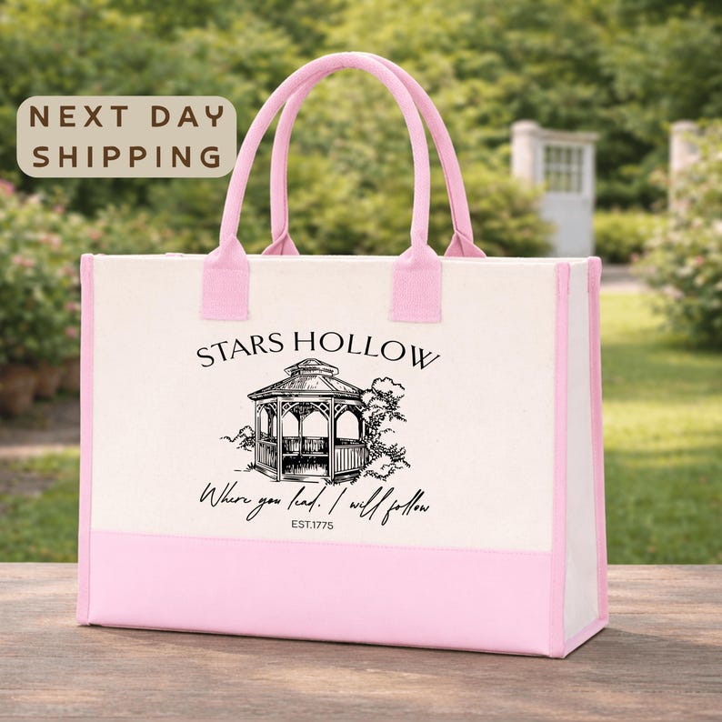 A pink tote bag with a printed design of a gazebo and the text "STARS HOLLOW" printed on it, placed on a wooden surface.