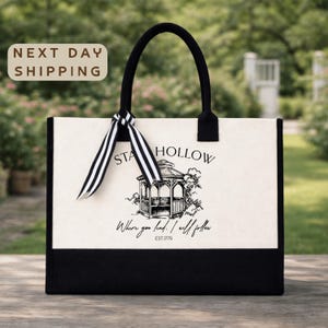 a black and white tote bag with a graphic design and text printed on it, placed on a wooden surface against a blurred background of greenery.