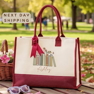 A red and white tote bag with a floral design and the name "Ashley" printed on it, placed on a wooden surface. There is a basket of flowers and a pair of sunglasses nearby.