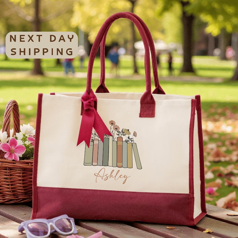 A red and white tote bag with a floral design and the name "Ashley" printed on it, placed on a wooden surface. There is a basket of flowers and a pair of sunglasses nearby.