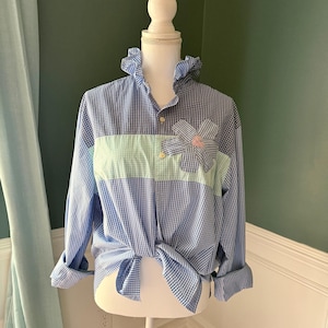May include: Blue and white gingham shirt with a light blue ruffle collar and a light green stripe across the chest. A fabric flower with a pink center is sewn on the chest. The sleeves are rolled up.
