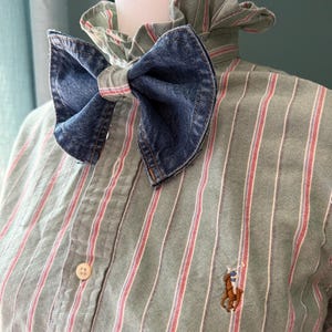 Könnte beinhalten: Eine Jeans-Schleife wird mit einem grün-rot gestreiften Button-Down-Hemd kombiniert. Das Hemd hat einen Rüschenkragen und ein kleines gesticktes Logo einer Person auf einem Pferd. Die Schleife ist aus blauem Jeansstoff.