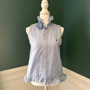 Op de afbeelding: Een lichtblauw mouwloos shirt met een gerimpelde kraag en zoom, weergegeven op een mannequin. Het shirt heeft een knoopsluiting aan de voorkant, een klein geborduurd logo en een decoratief ruitaccent bij de kraag.