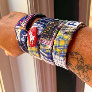 May include: A set of stacked fabric bracelets featuring various plaid and checkered patterns in blue, yellow, and white. The bracelets include embroidered patches with the text Polo Ralph Lauren and a red leather-like horse logo emblem.