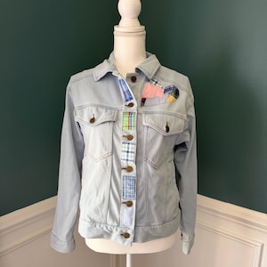 May include: Light blue denim jacket with patchwork accents. The jacket features a classic design with button closures and pockets. Colorful fabric patches are sewn on the front, adding a unique, handcrafted touch. The jacket is displayed on a mannequin.