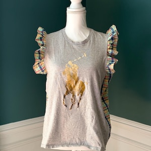May include: A grey sleeveless top featuring a large gold metallic polo player logo on the front. The garment has ruffled armholes and side panels made of a multi-colored plaid fabric in shades of blue, yellow, green, and red, with a round neckline.