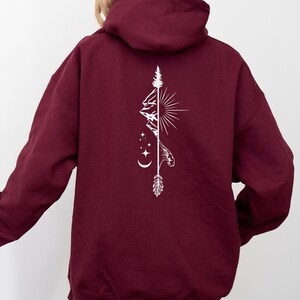 May include: Burgundy hoodie with a white graphic on the back. The graphic features a mountain range, sun, stars, moon, and an arrow. The hoodie has a hood and long sleeves.