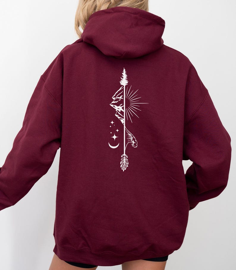 May include: Burgundy hoodie with a white graphic on the back. The graphic features a mountain range, sun, stars, moon, and an arrow. The hoodie has a hood and long sleeves.