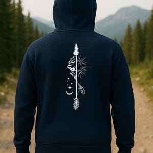 May include: Navy blue hoodie with a white graphic on the back featuring a mountain range, sun, arrow, and stars. The hoodie has a hood and a drawstring. The background shows a forest and a mountain range.