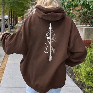 May include: A brown hoodie with a white graphic design on the back. The design features a mountain, sunburst, arrow, and moon and stars. The hoodie is a pullover style with a hood.