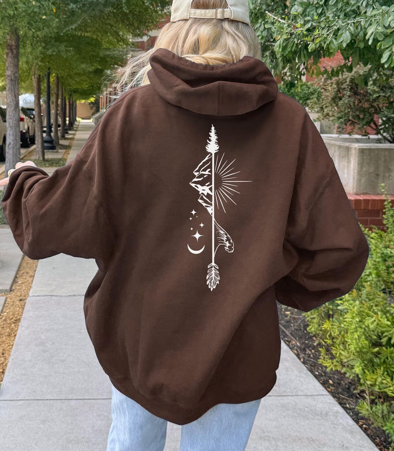 May include: A brown hoodie with a white graphic design on the back. The design features a mountain, sunburst, arrow, and moon and stars. The hoodie is a pullover style with a hood.