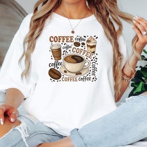 May include: White t-shirt with a coffee-themed design. The graphic includes coffee cups, beans, and the word "COFFEE" in different fonts. A casual, everyday wear item.