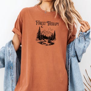 May include: Rust-colored t-shirt with the text "Forest Therapy" above a black graphic of a mountain scene with trees, a river, and a crescent moon. The shirt has rolled-up sleeves and is paired with a denim jacket.