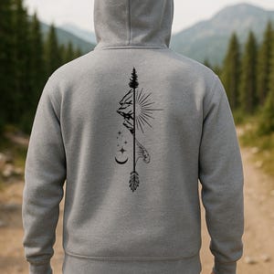 May include: A gray hooded sweatshirt with a black graphic design on the back. The design features a mountain, sun, stars, moon, and feather. The sweatshirt has a drawstring hood and long sleeves. The background shows a forest and mountains.
