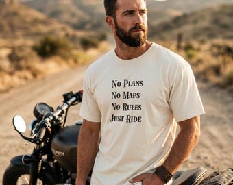 May include: Cream-colored t-shirt with the text "No Plans, No Maps, No Rules, Just Ride" in black. A black motorcycle is visible in the background. The shirt is short-sleeved.