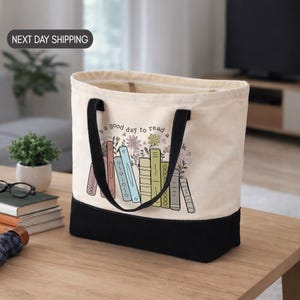 May include: A canvas tote bag with a black base and handles. The bag features a design of colorful books and flowers with the text "s a good day to read a k". The bag is sitting on a wooden table.