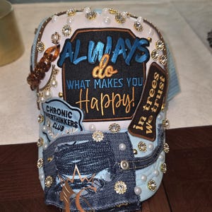May include: A light blue and denim baseball cap with the text "ALWAYS do WHAT MAKES YOU Happy!" embroidered on the front. The cap is embellished with pearls, rhinestones, and a brown beaded bracelet.