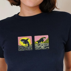 Puede incluir: Camiseta azul marino con dos gráficos cuadrados de orcas sobre un fondo rosa y amarillo. La palabra "black" está escrita en cursiva en la parte inferior izquierda. Prenda de vestir informal.