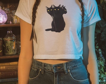 May include: A white short-sleeved crop top featuring a graphic print of a black cat with three heads and bright yellow eyes. The shirt is paired with dark blue denim jeans that have a button and zipper closure at the waist.