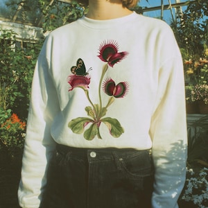 May include: A white long-sleeve crewneck sweatshirt featuring a botanical illustration of three Venus flytrap plants with green leaves and dark red traps, with a small butterfly perched on one of the traps, centered on the chest area.