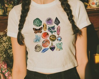 May include: A white short-sleeved t-shirt featuring a central graphic print of various colorful crystals, gemstones, a butterfly, a small plant, and a fossil. The design includes diverse shapes and textures in shades of green, purple, blue, and gold.